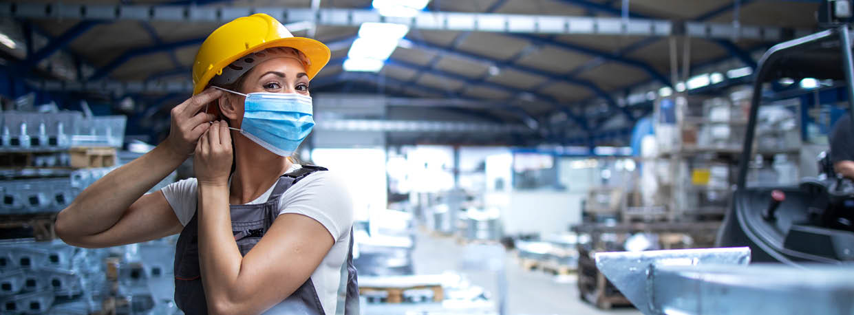 Shot of female factory worker in uniform and hardhat putting on face mask in industrial production plant  People working during COVID-19 pandemic 