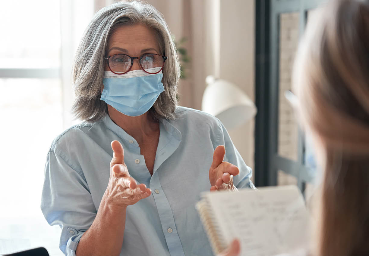 Old middle aged female mentor, teacher, hr manager wearing face mask training young worker intern, teaching student, interviewing job seeker at meeting in office  Social distance and safety at work 