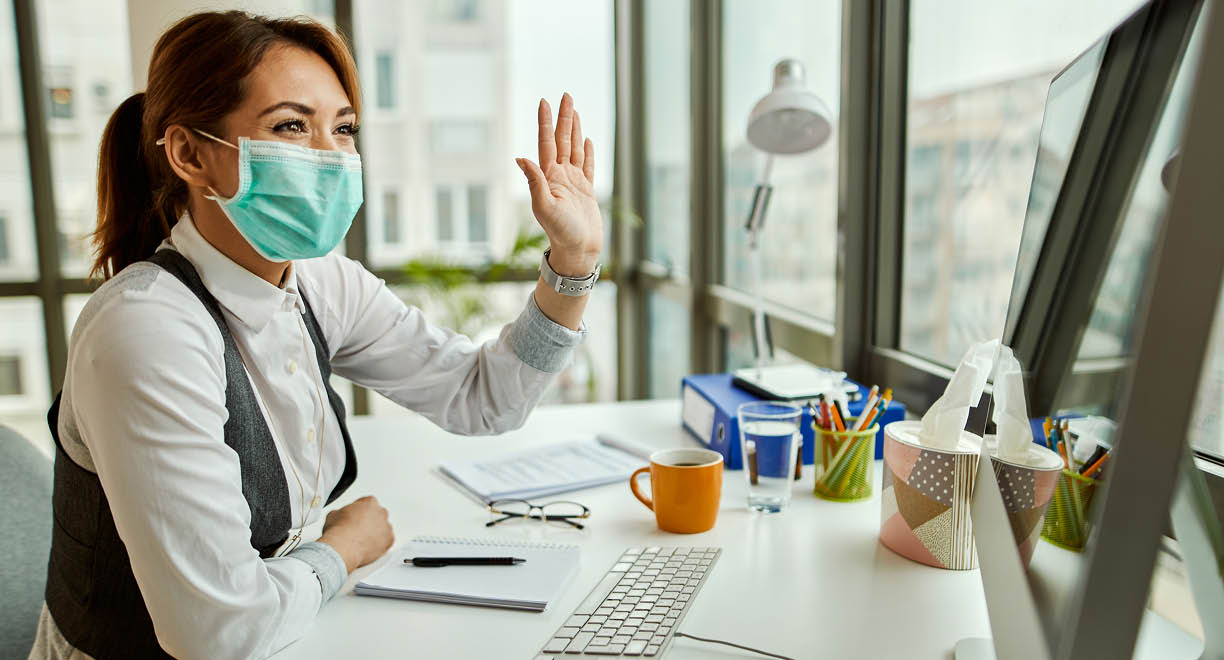 Happy businesswoman with face mask using computer and waving to someone while making video call from her office  