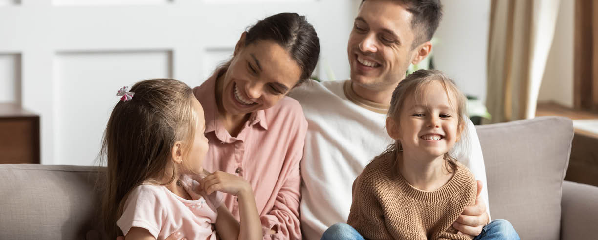 Happy young mother and father with two little daughters sitting on couch, looking at each other, family enjoying tender moment, smiling parents and preschool children having fun together