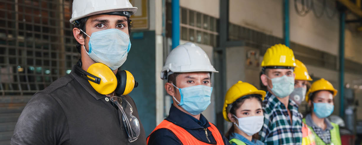 Group of diverse team of workers wearing face mask and protective helmets standing in front of the factory