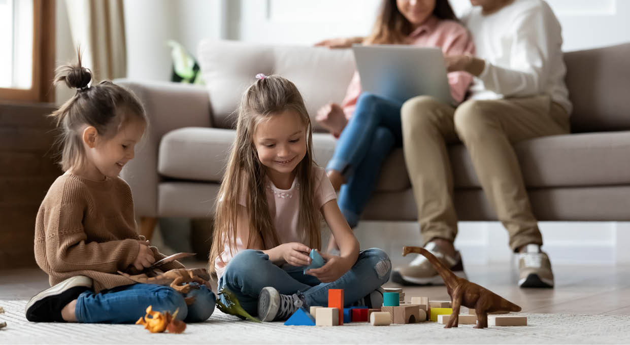Happy little kids siblings preschool daughters having fun playing toys on floor while parents sit on sofa using laptop in comfy living room, family relaxing at home enjoying cozy leisure lifestyle