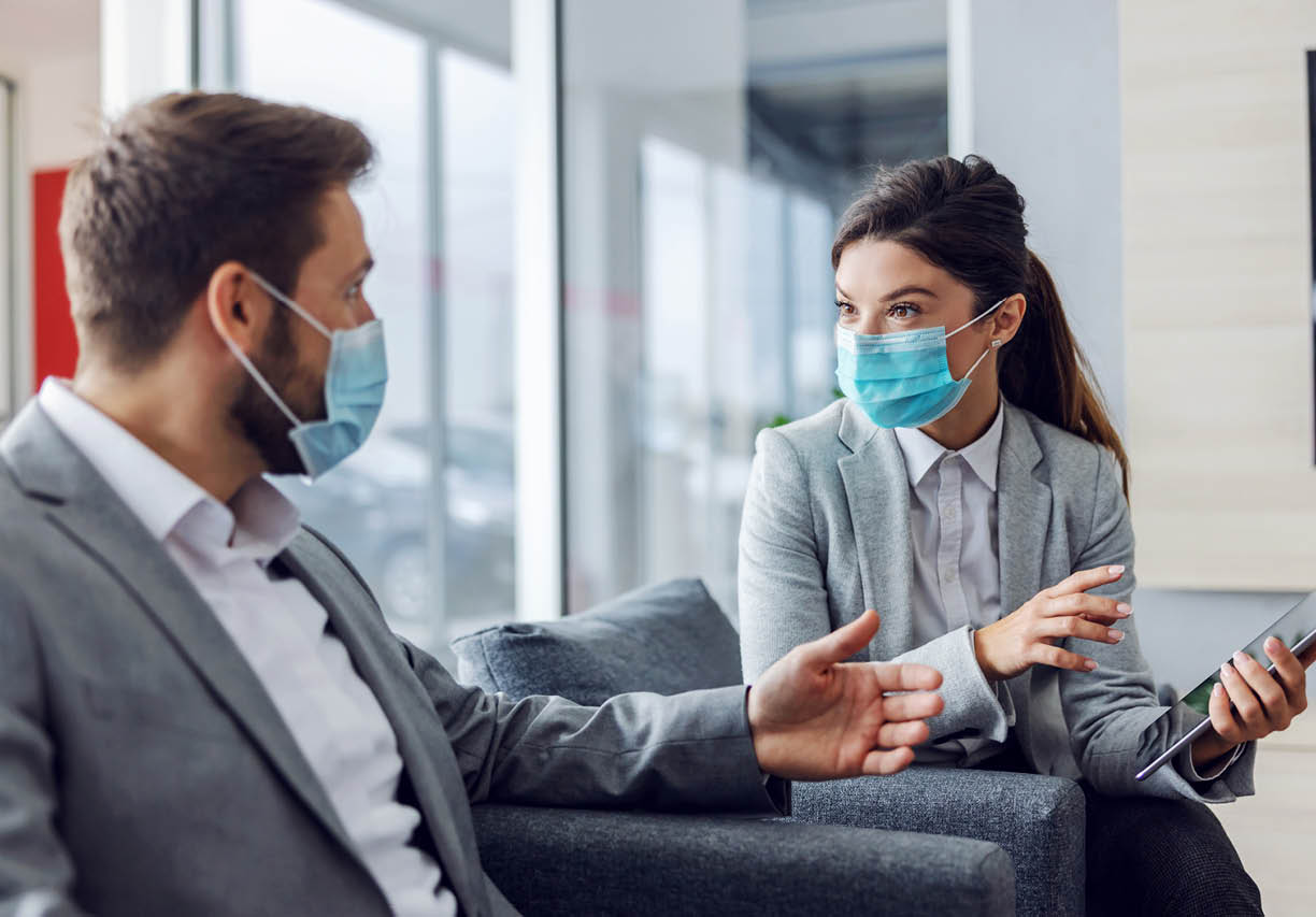 Friendly car seller with face mask sitting with customer in car salon, holding tablet and talking about specifications and performances of car during corona virus 