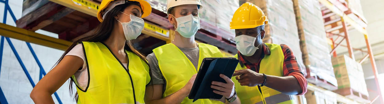 Warehouse workers team meeting using tablet computer with wearing face mask and protective safety helmets standing in storage factory standard to prevent coronavirus COVID-19 infection